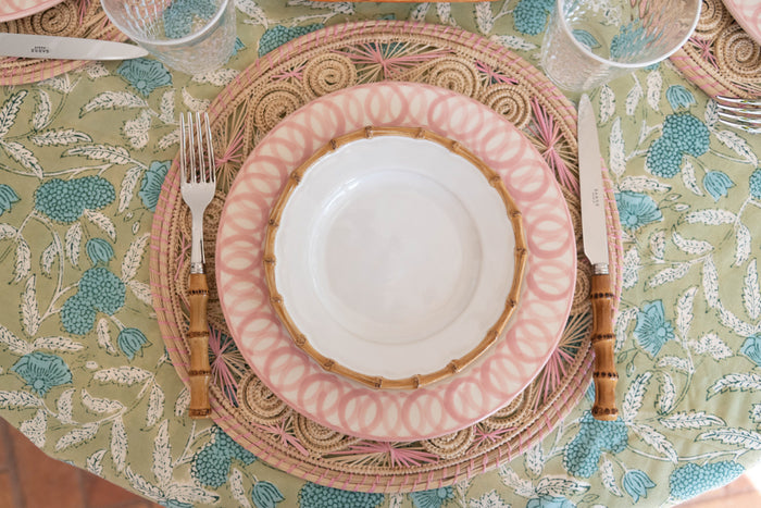 Round natural bamboo cocktail plate under pink patterned dinner plate and white bowl on green floral tablecloth with.