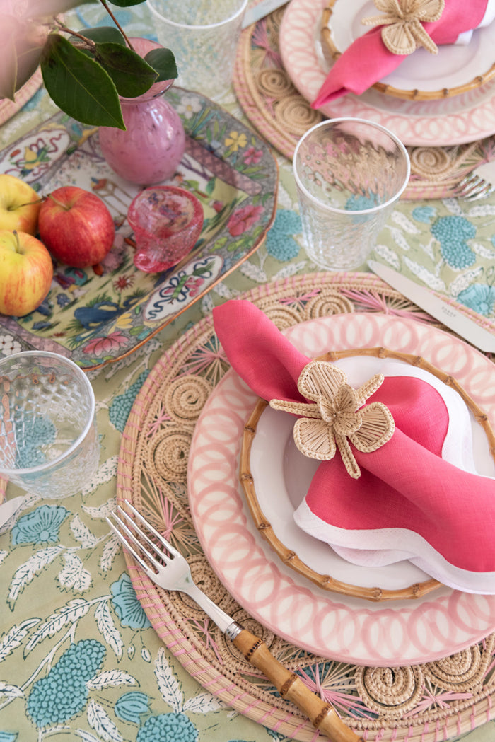 Round bamboo cocktail plate with woven pattern under pink and white layered dinnerware and bright pink napkin with flower.