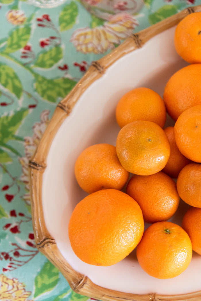 Oval bamboo serving platter with natural finish holding bright fresh oranges on patterned tablecloth.