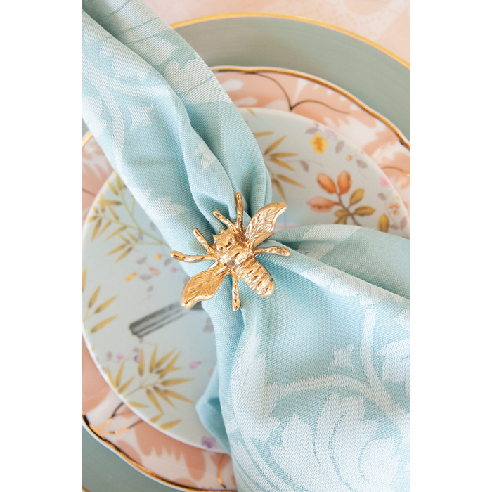Gold bee-shaped napkin ring securing a light blue patterned cloth napkin on floral and pastel dinner plates.