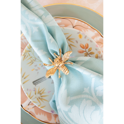 Gold bee-shaped napkin ring securing a light blue patterned cloth napkin on floral and pastel dinner plates.
