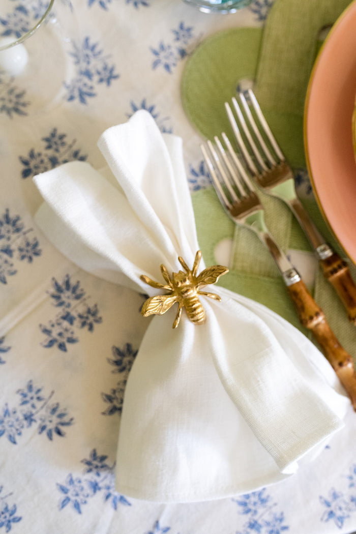 Gold bee-shaped napkin ring holding a white cloth napkin on a blue floral tablecloth next to green napkin and brown-handled.