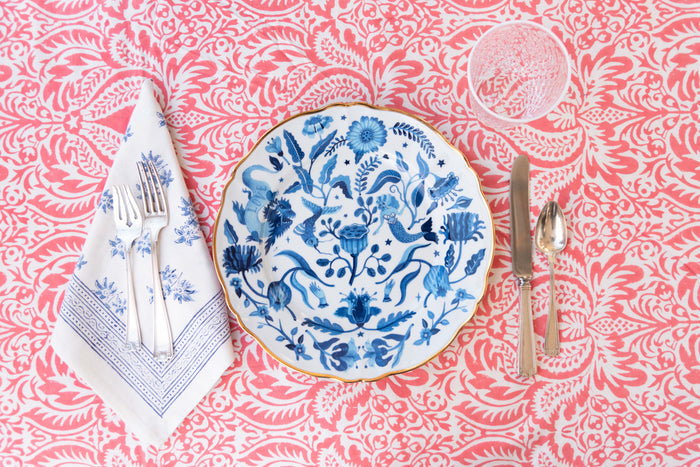 Blue and white ceramic dinner plate with intricate floral and bird pattern on a red patterned tablecloth with silver cutlery .