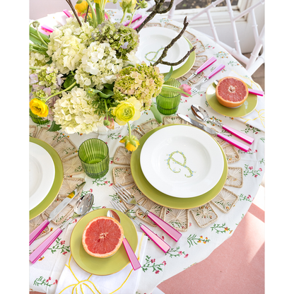 White dinner plate with green rim and decorative green monogram, set on floral tablecloth with pink handles flatware and.