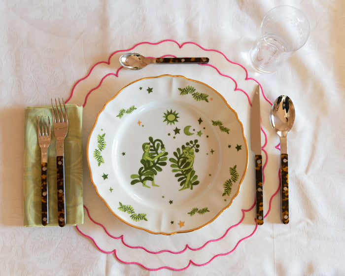 White ceramic dinner plate with green botanical designs, placed on scalloped pink-edged placemat with gold and black cutlery .