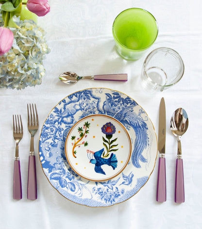 Small white plate with colorful floral and bird design, placed on larger blue and white patterned plate with purple-handled.