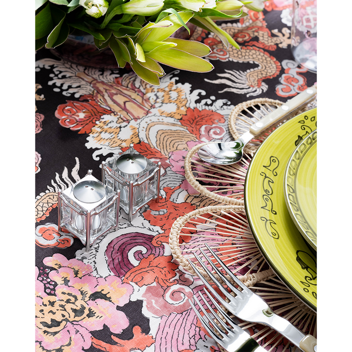 A close-up of a vibrant tablescape featuring a bold multicolored dragon-print tablecloth, woven floral placemat, lime green patterned plates, and pearl-handled flatware. Two small glass candle holders and a lush green floral arrangement add decorative detail to the setting.