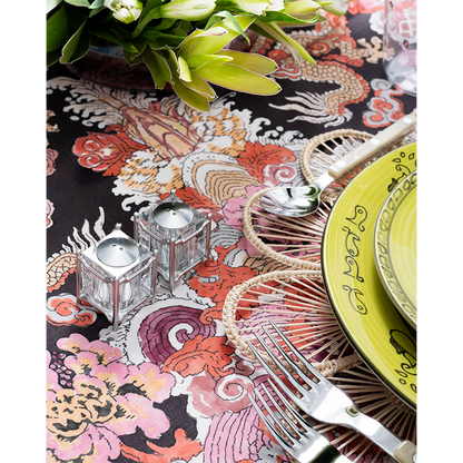 A close-up of a vibrant tablescape featuring a bold multicolored dragon-print tablecloth, woven floral placemat, lime green patterned plates, and pearl-handled flatware. Two small glass candle holders and a lush green floral arrangement add decorative detail to the setting.