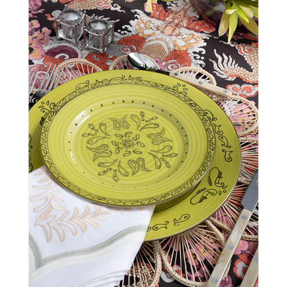A vibrant place setting featuring a lime green patterned plate layered over a woven floral-shaped placemat and embroidered white napkin. Pearl-handled flatware, pink glassware, and a bold multicolored dragon-print tablecloth complete the playful tablescape.