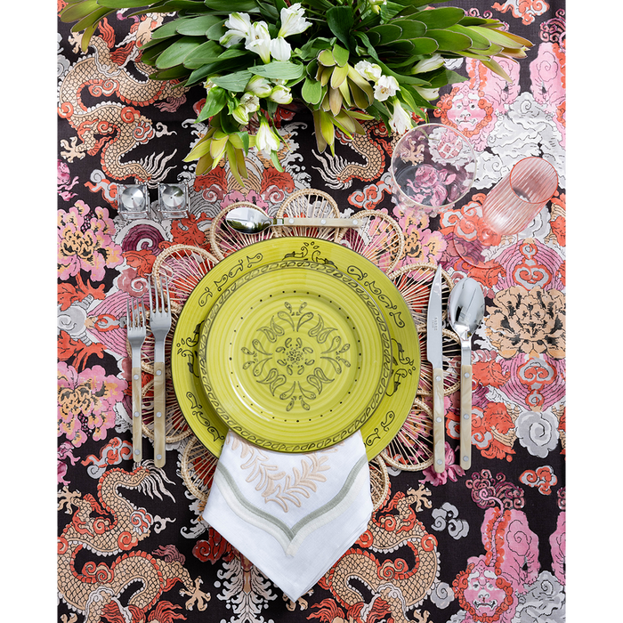 A vibrant tablescape featuring a lime green patterned plate layered over a woven placemat and embroidered white napkin. Colorful dragon-print fabric, polished flatware, pink glassware, and a lush arrangement of white flowers create a bold, playful setting.