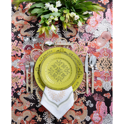 A vibrant tablescape featuring a lime green patterned plate layered over a woven placemat and embroidered white napkin. Colorful dragon-print fabric, polished flatware, pink glassware, and a lush arrangement of white flowers create a bold, playful setting.