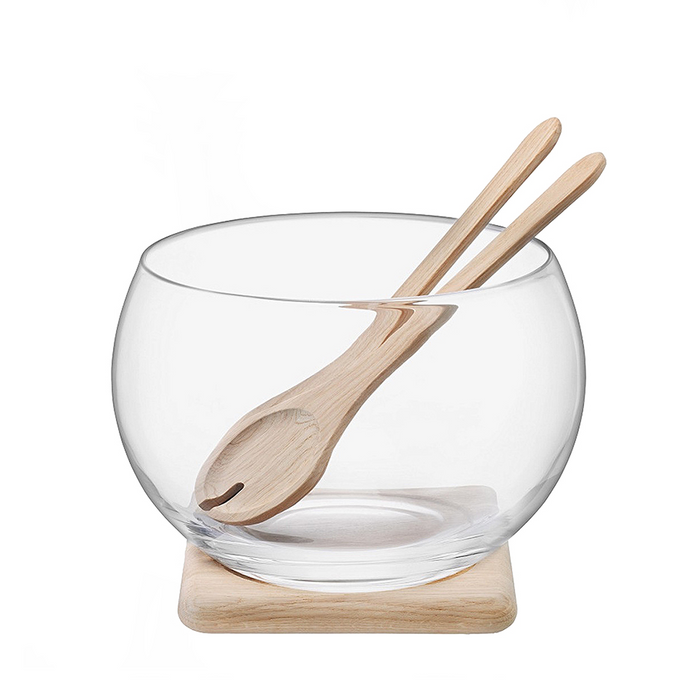 Clear glass salad bowl with rounded shape, wooden salad tongs, and square wooden base.