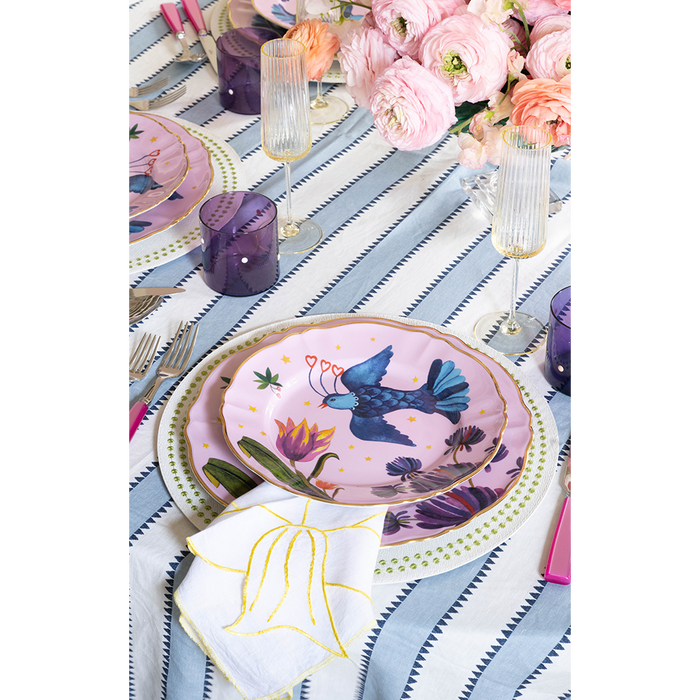 Chartreuse placemat with blue bird and floral print under pink and white plates, set on striped tablecloth.