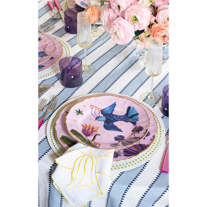 Chartreuse placemat with blue bird and floral print under pink and white plates, set on striped tablecloth.