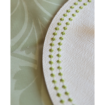 Round white placemat with textured fabric and chartreuse dotted stitched border on green floral tablecloth.