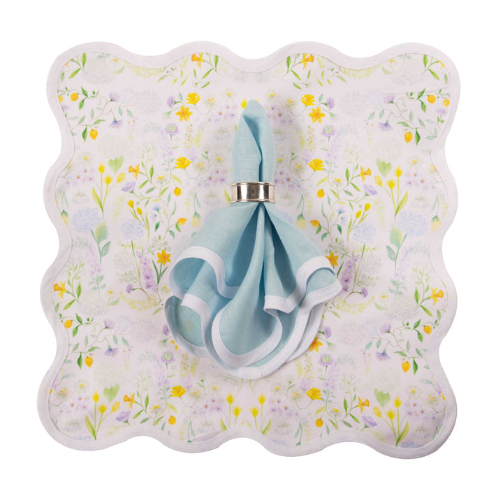 Sky blue linen napkin with scalloped edges on a white floral print napkin with yellow and purple flowers.