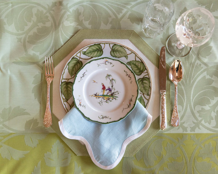 Sky blue linen napkin with scalloped edges folded beneath floral china on pale green damask tablecloth with gold flatware.