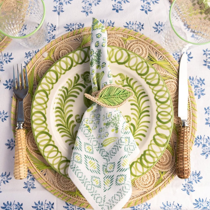 Woven rattan napkin ring shaped like a green ivy leaf around patterned cloth napkin on leafy plate.
